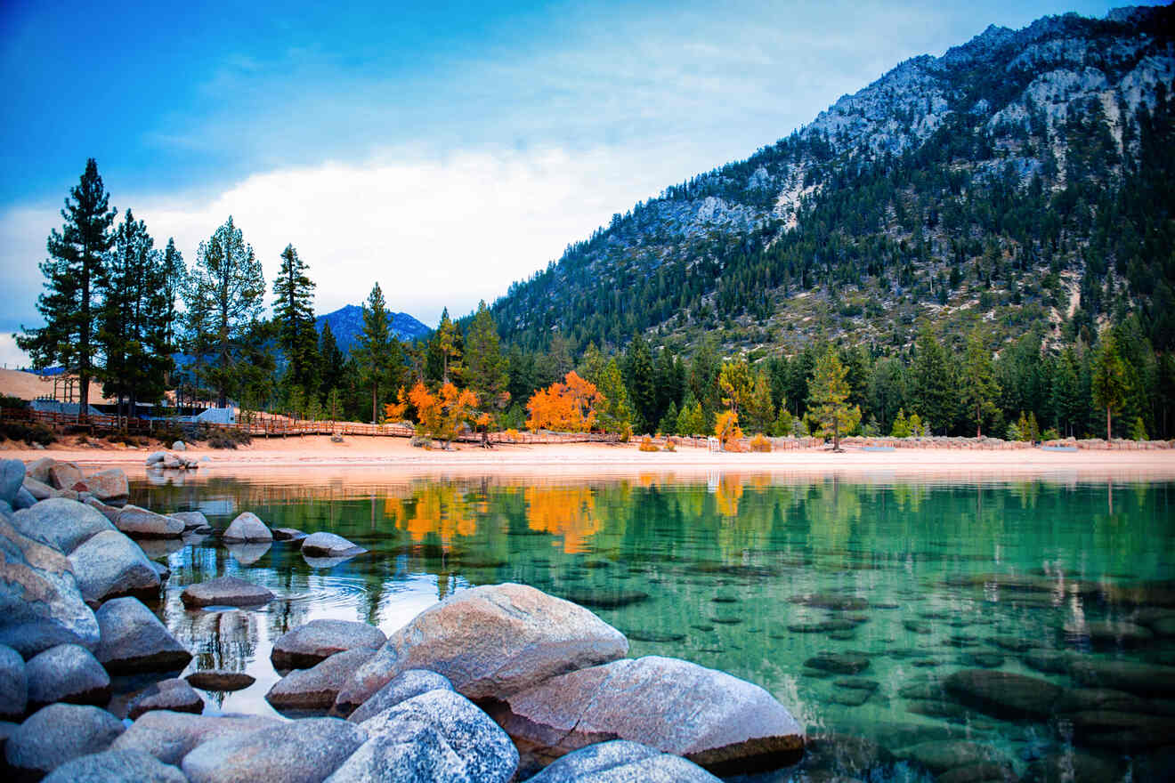 Serene lake view with clear waters, autumnal trees on the shore, and a backdrop of mountains under a soft blue sky.